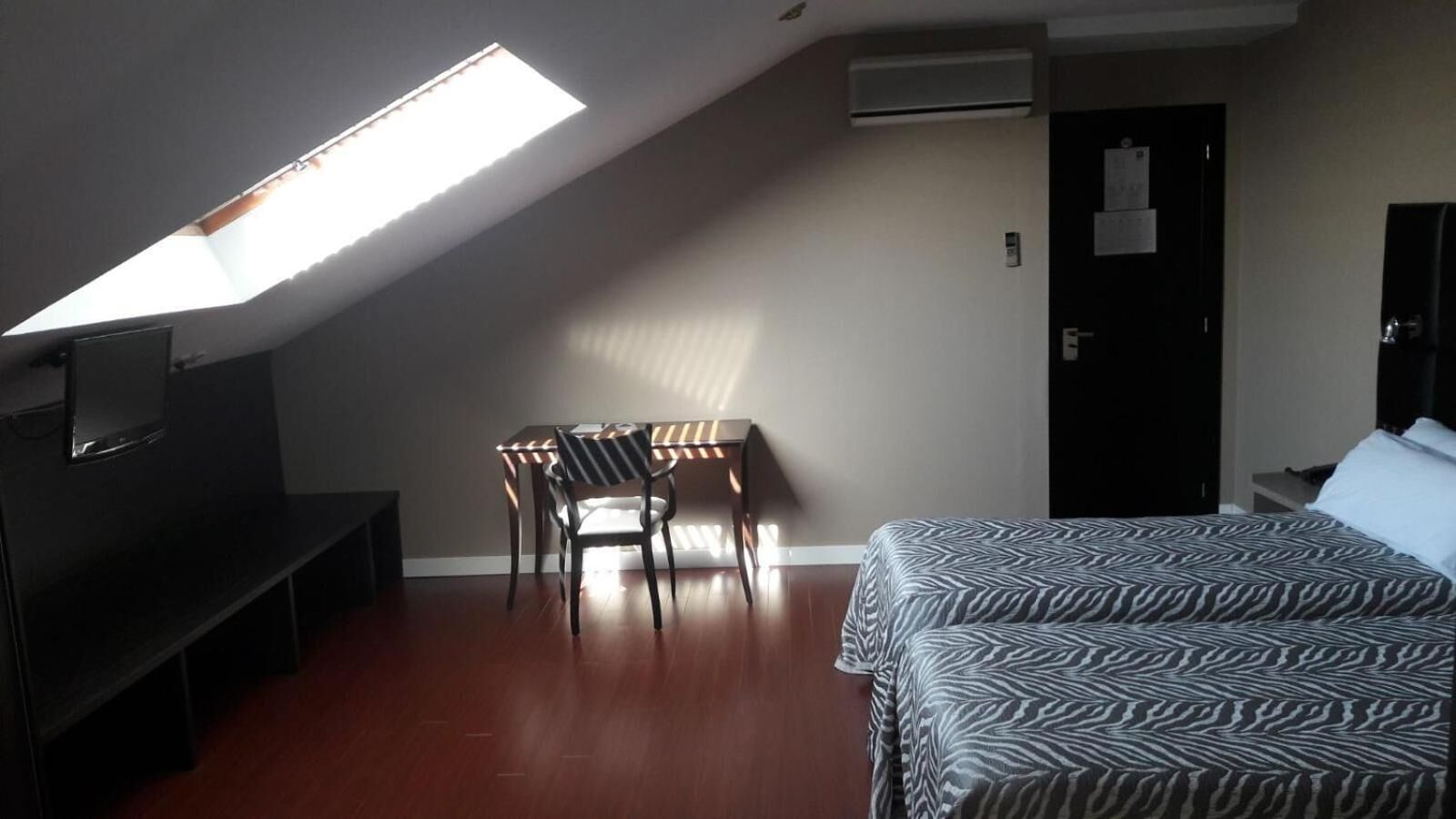 A room with a bed, a desk with a chair, a TV, and a skylight.