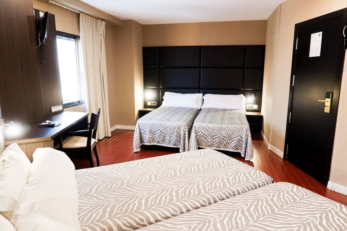 A hotel room with two beds, a desk, a chair, a TV, and a door.