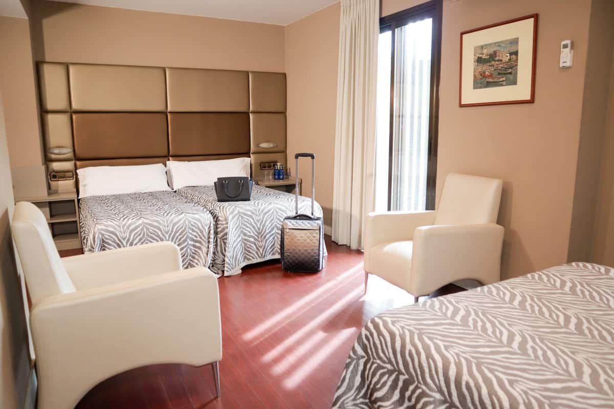 A hotel room with two beds, a sitting area, a window with curtains, and a painting on the wall.