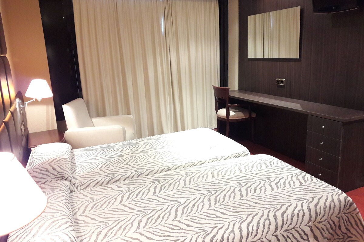 A hotel room with a bed, a desk, a chair, a mirror, and a window with curtains.