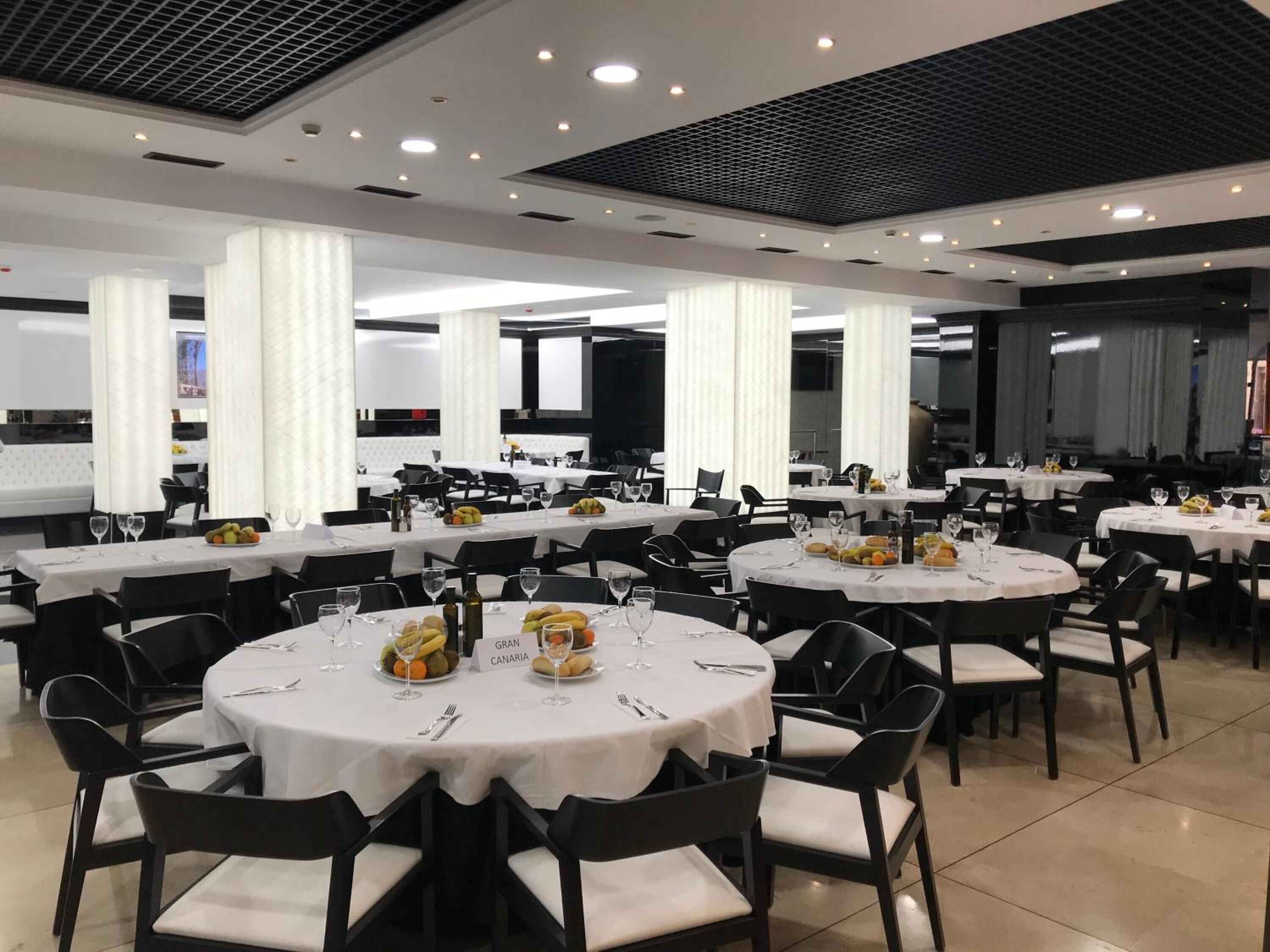 A spacious dining area with multiple round tables, each set with white tablecloths, black chairs, and a variety of fruits and bottles.