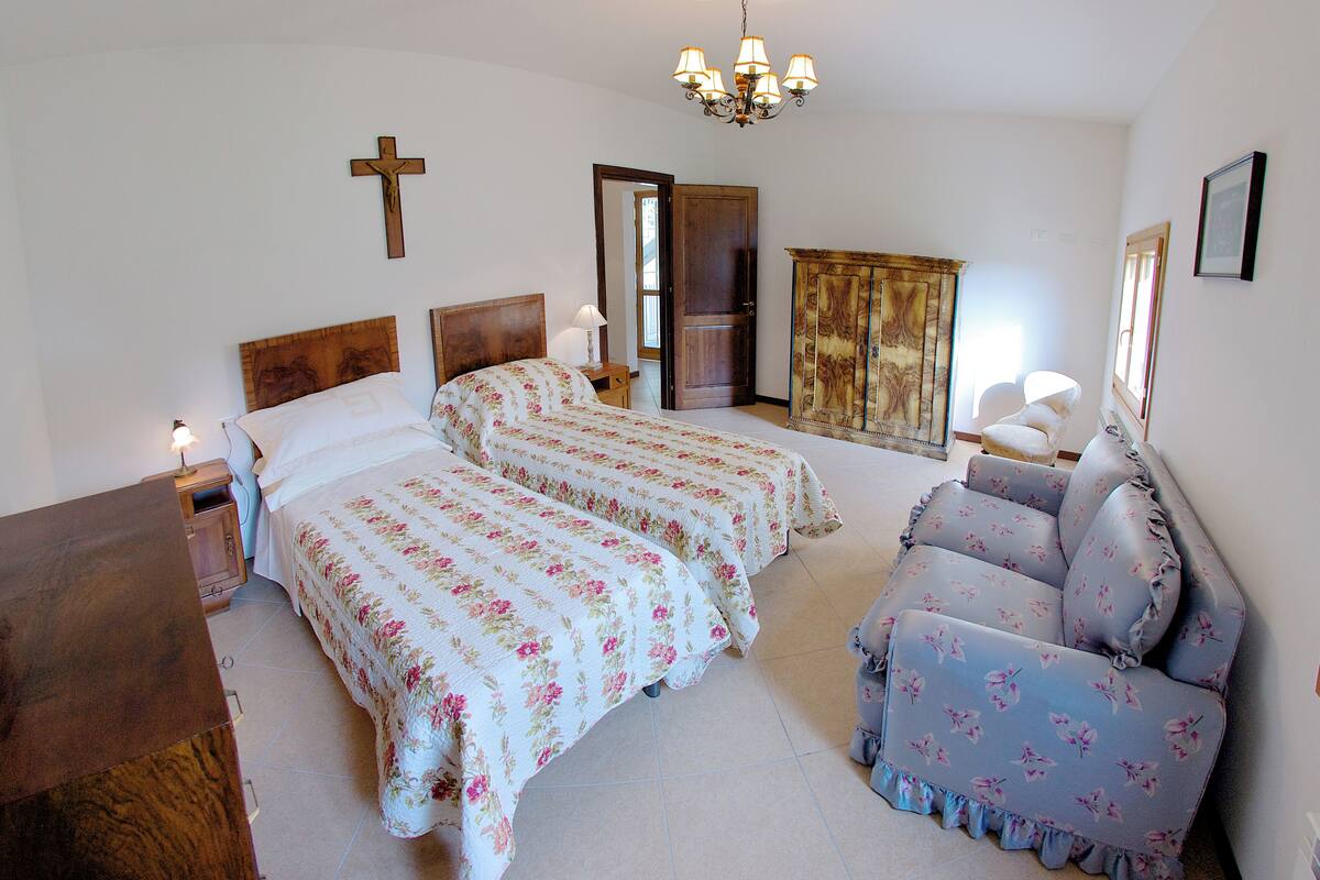 A bedroom with two beds, a sofa, a wooden cabinet, a cross on the wall, and a chandelier.