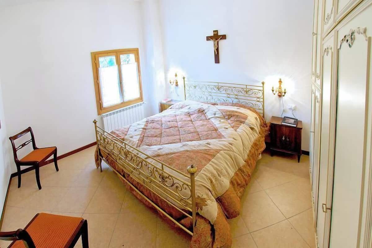 A bedroom with a metal bed frame, a wooden chair, a bedside table, a window, and a wall-mounted cross.