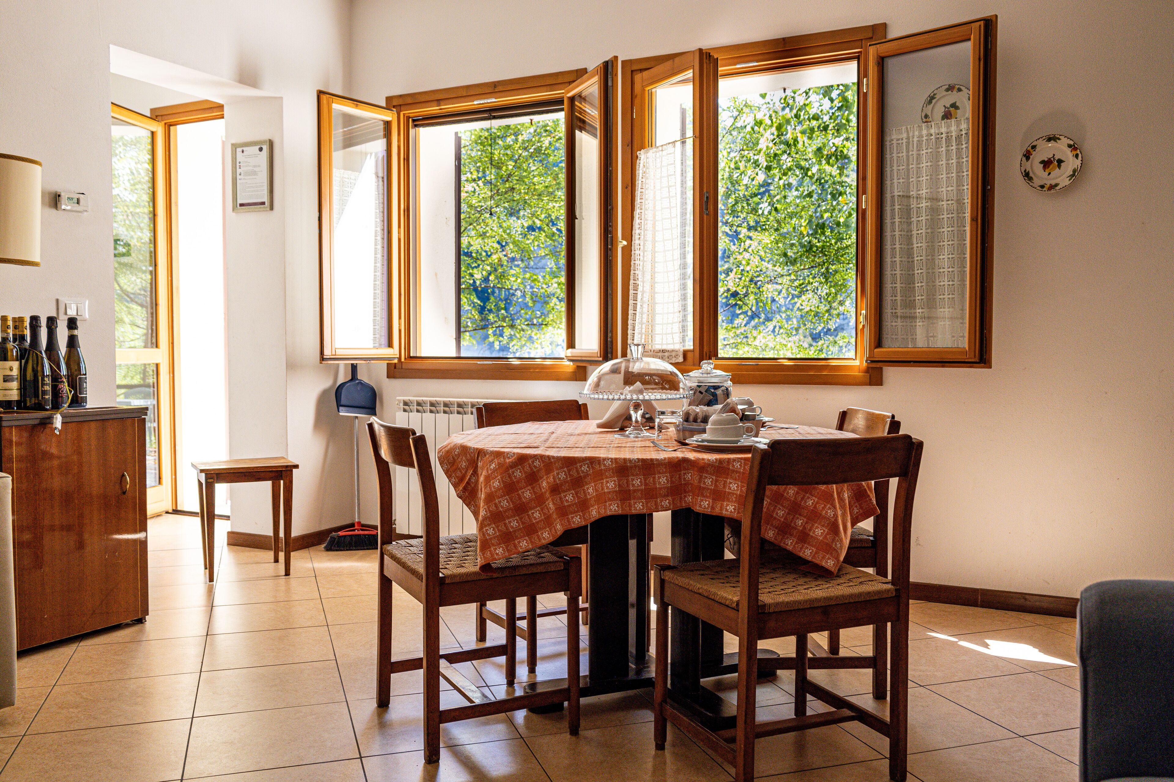 A dining area with a table set for four, surrounded by wooden chairs, a wooden cabinet, and a window with a view of trees.