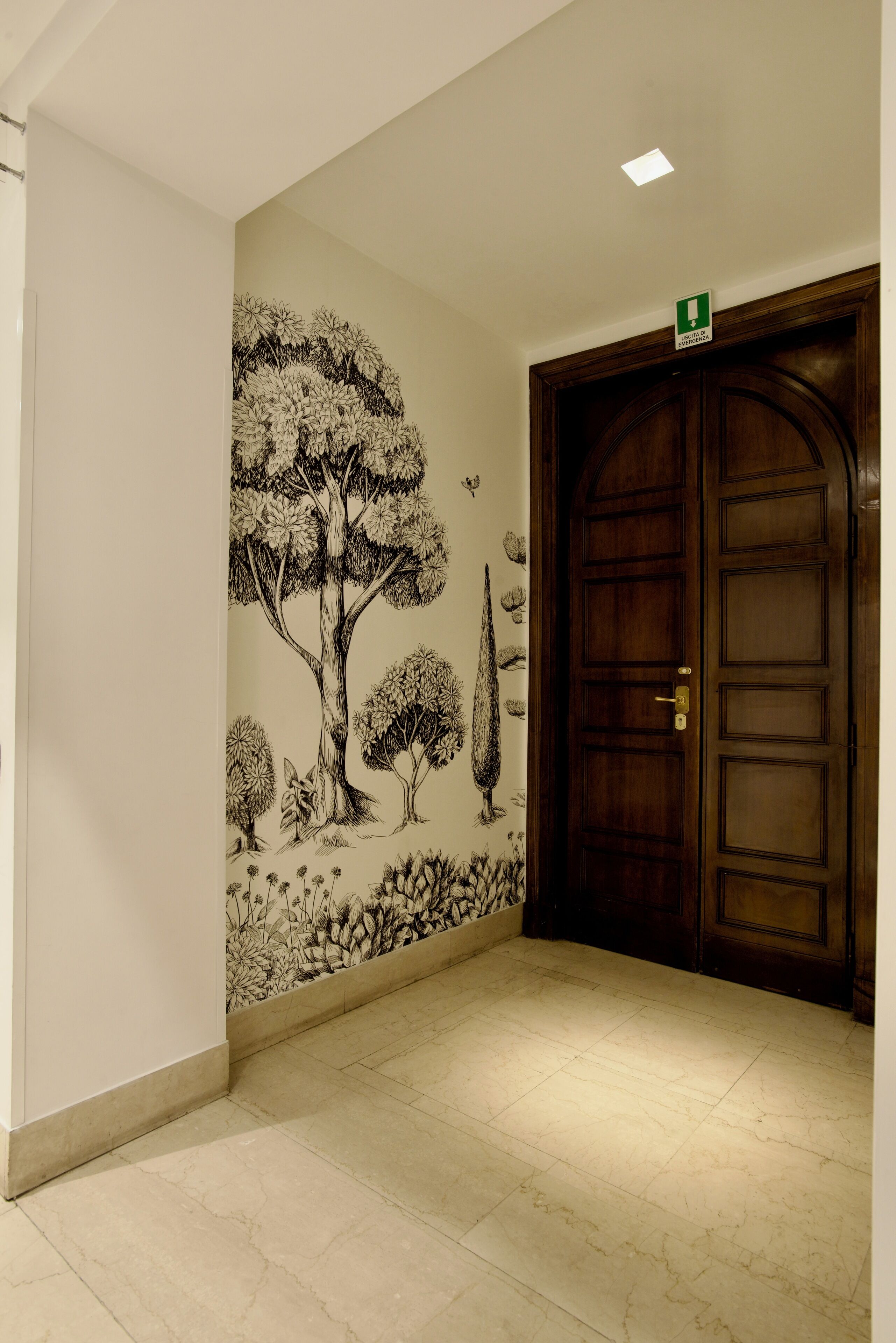 A hallway with a wooden door, a green emergency exit sign, and a wall mural of trees and plants.
