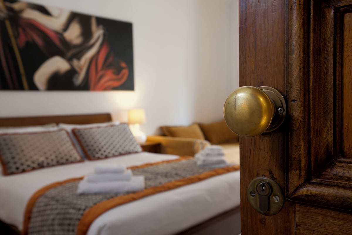 A hotel room with a wooden door, a bed with white bedding and a patterned throw, and a painting on the wall.
