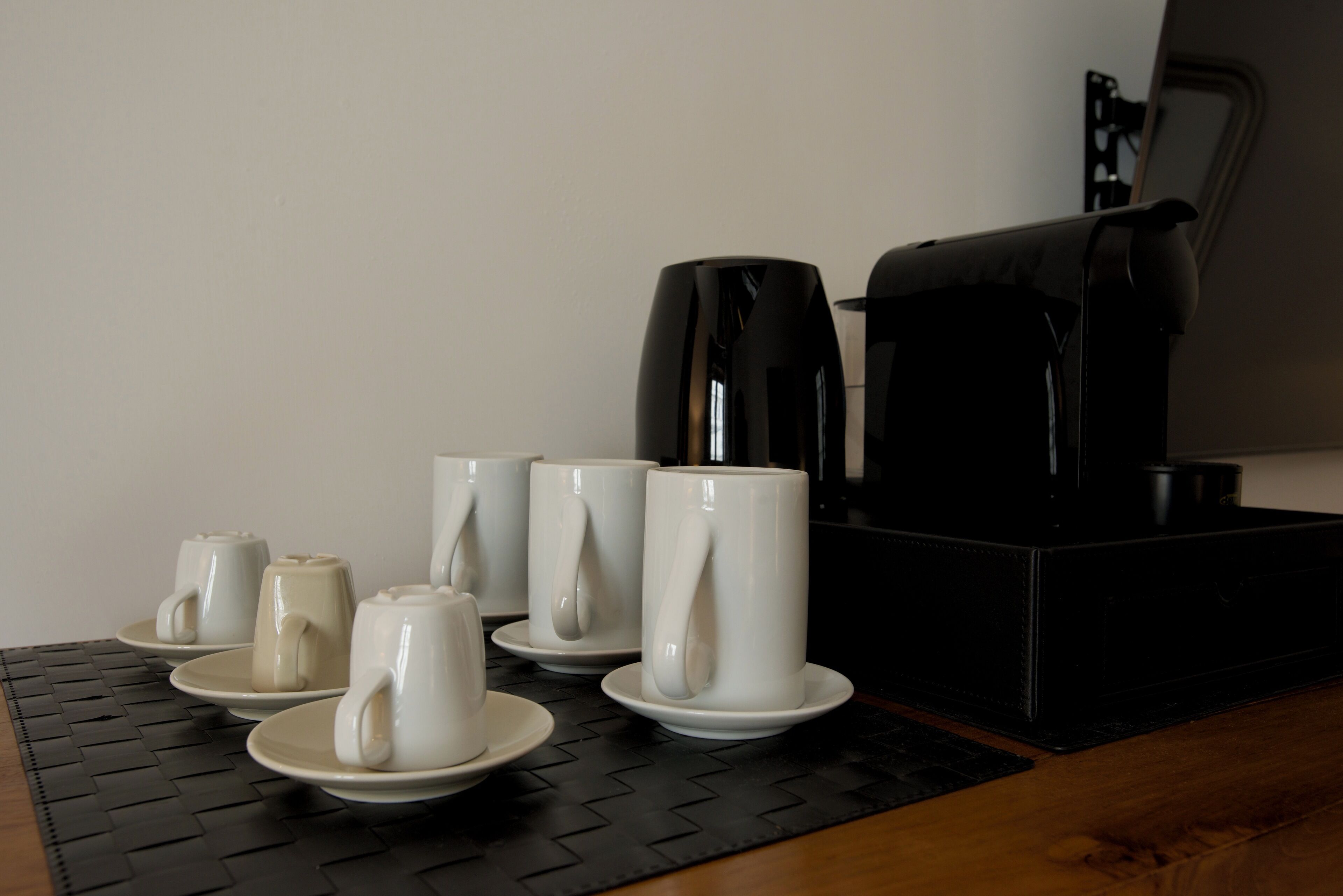 A coffee station with multiple white mugs and saucers, a black coffee maker, and a black tray on a wooden table.