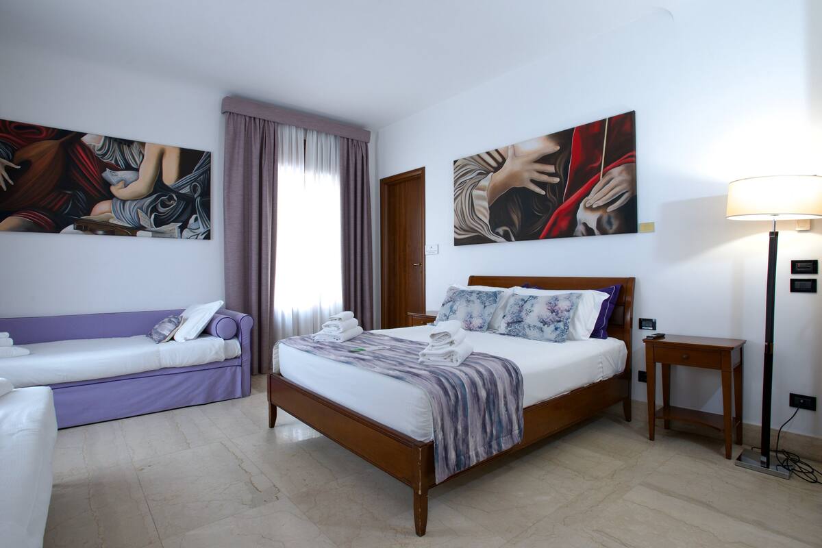 A hotel room with a bed, a sofa, and two paintings on the wall.