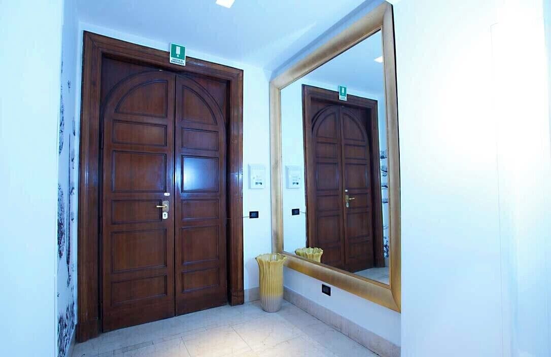 A hallway with two wooden doors, each marked with a green sign, and a large mirror reflecting the doors.