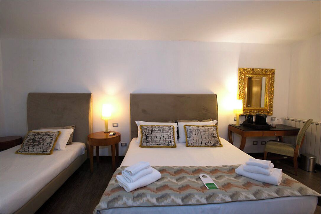 Ein Hotelzimmer mit zwei Einzelbetten, einem Kingsize-Bett, einem Schreibtisch mit Stuhl, einem Spiegel und je eine Nachttischlampe neben dem Kingsize-Bett.