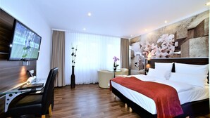 A hotel room with a bed, a desk with a chair, a TV, and a small table with a vase. - Ascot Hotel (Remscheid)