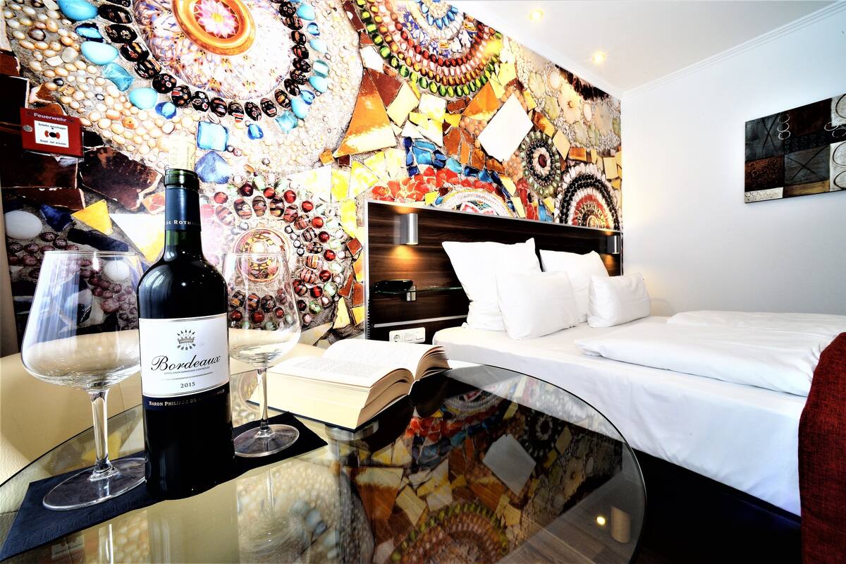 A hotel room with a bed, a glass table with a bottle of Bordeaux wine, and a book.