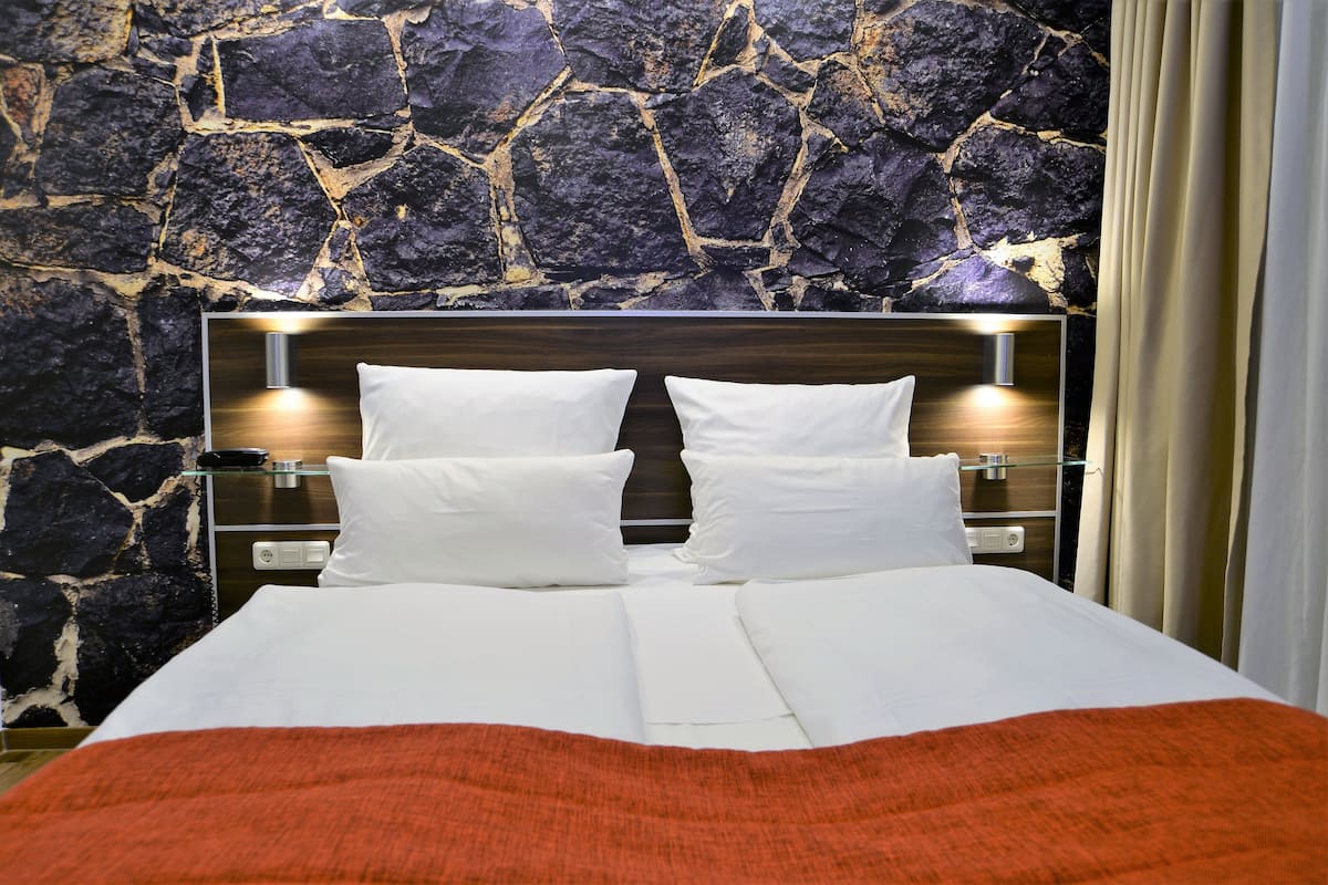 A neatly made bed with white linens and a red blanket, a wooden headboard, and a stone wall behind.