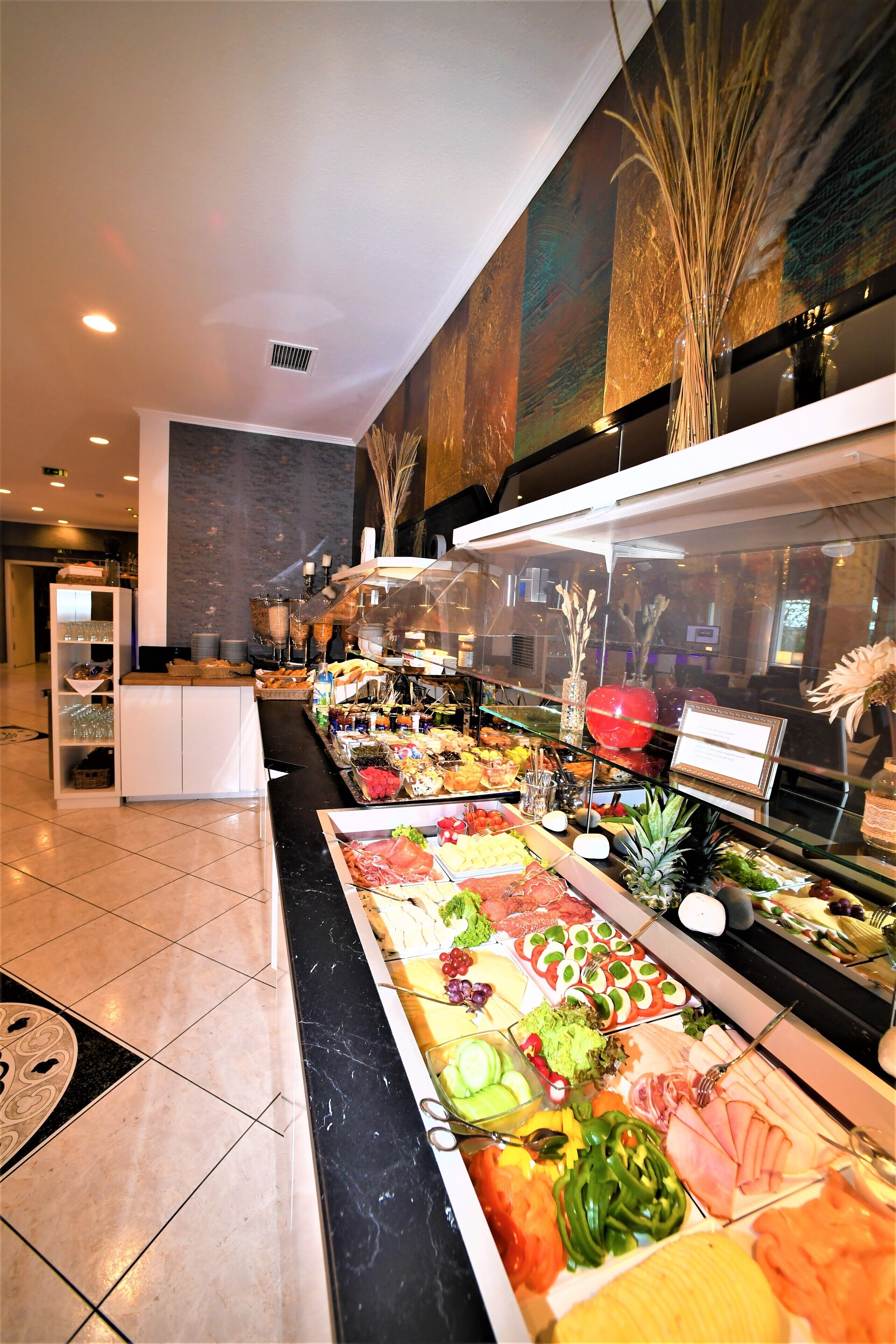 Un buffet con diverse portate, tra cui frutta, verdura e carni, in una sala ben illuminata con pavimento piastrellato e un grande quadro appeso al muro.
