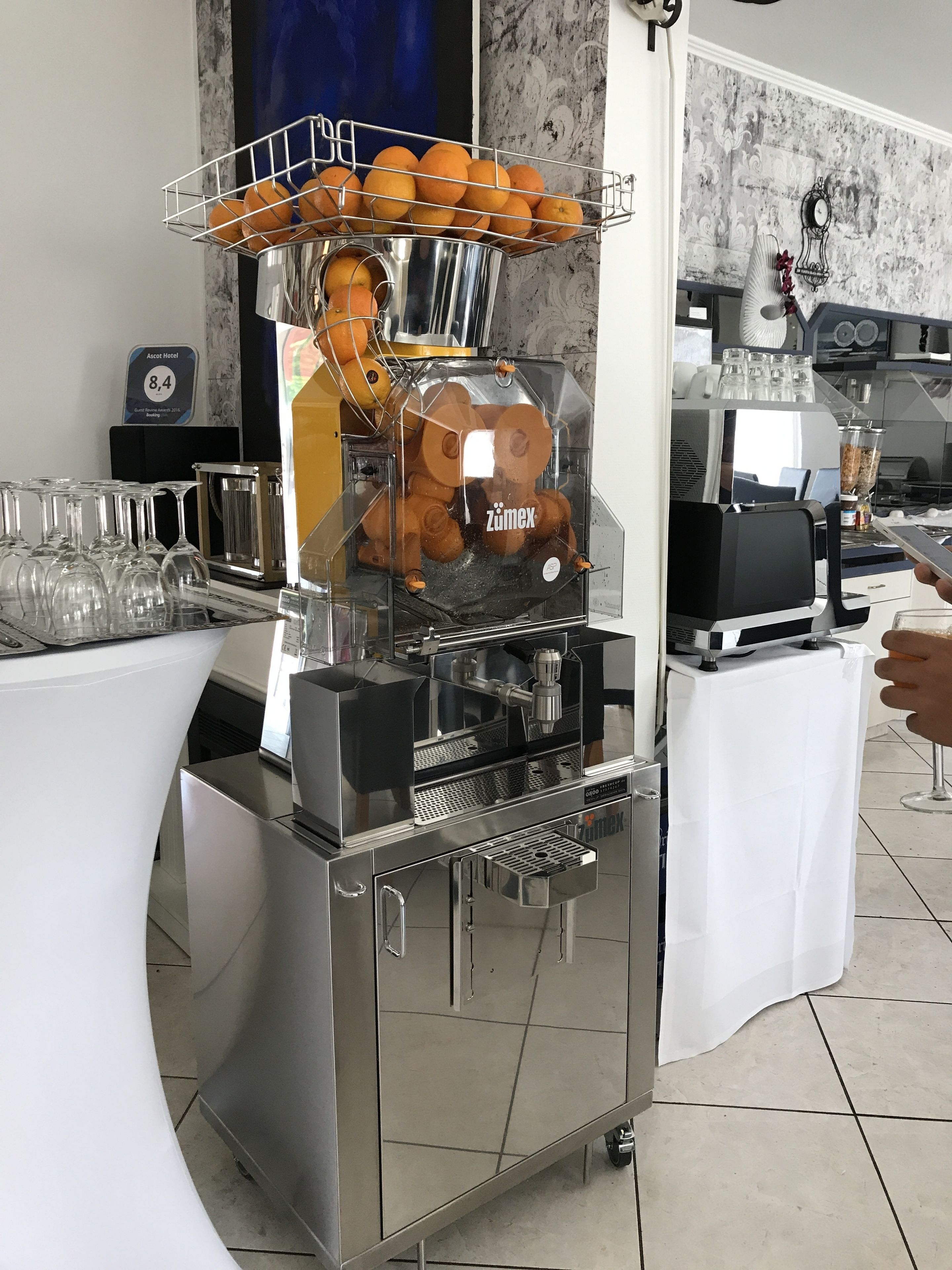 Una estación de café autoservicio con una máquina de café con la marca, un cesto de naranjas y una vitrina de vidrio.