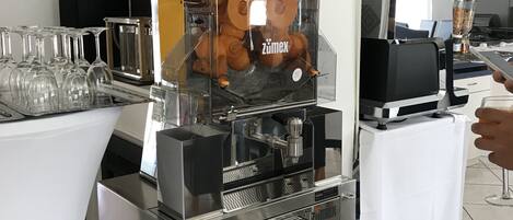 Una estación de café autoservicio con una máquina de café con la marca, un cesto de naranjas y una vitrina de vidrio.