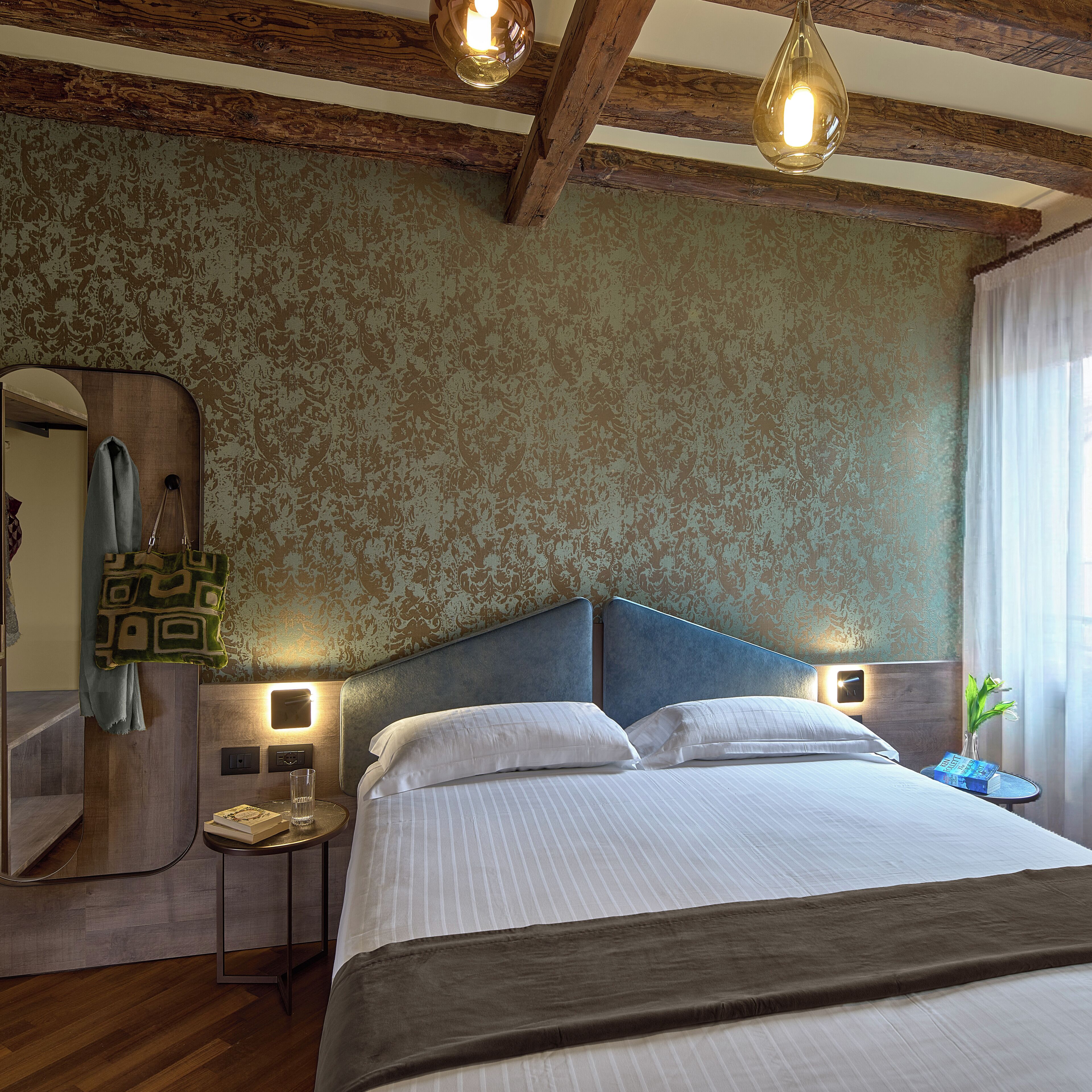 Quarto de hotel com uma cama grande, vigas de madeira no teto, um espelho e uma mesa de cabeceira com um livro e um copo.