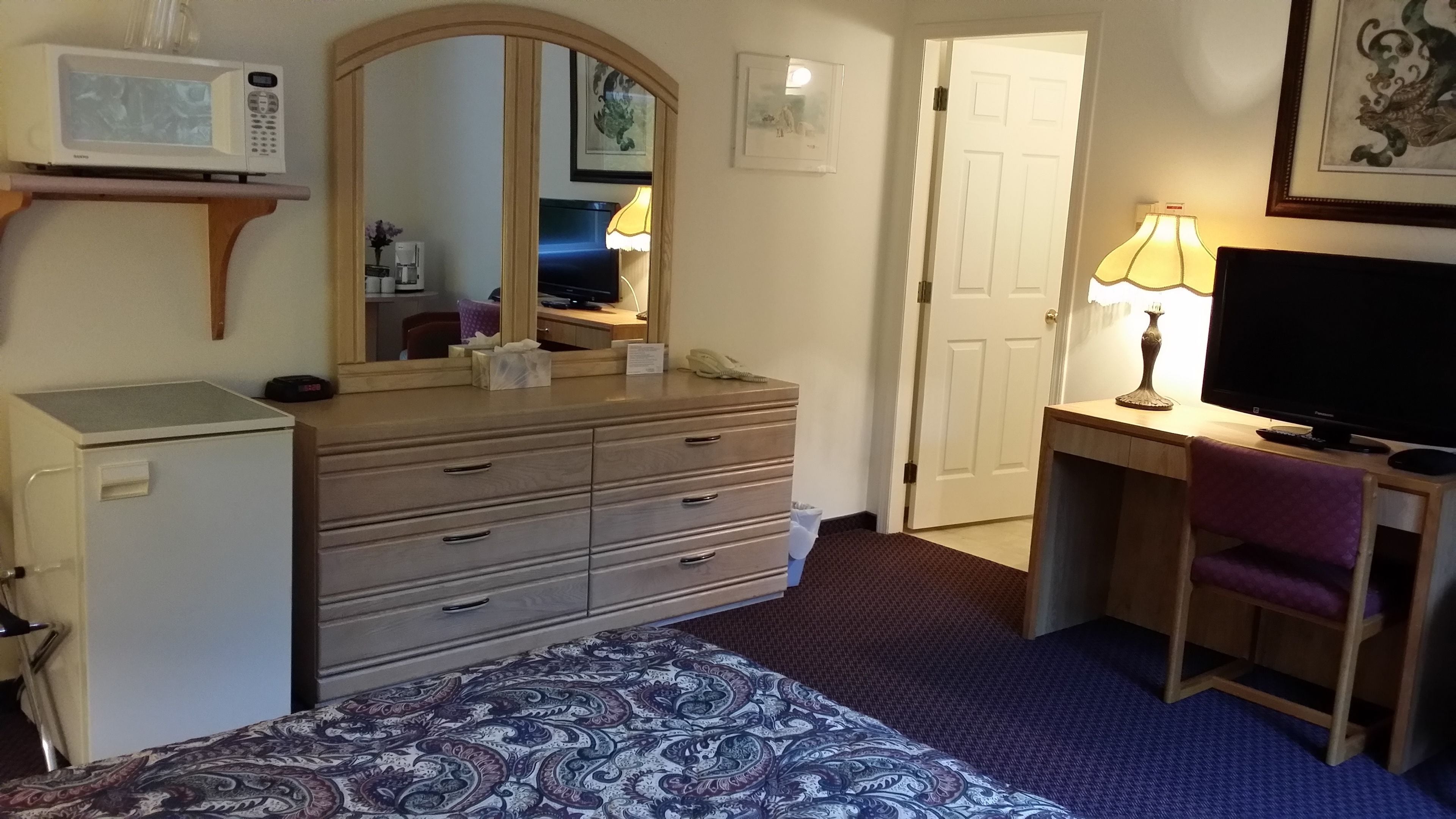 A hotel room with a bed, a dresser, a desk with a computer, a lamp, a TV, and a microwave.