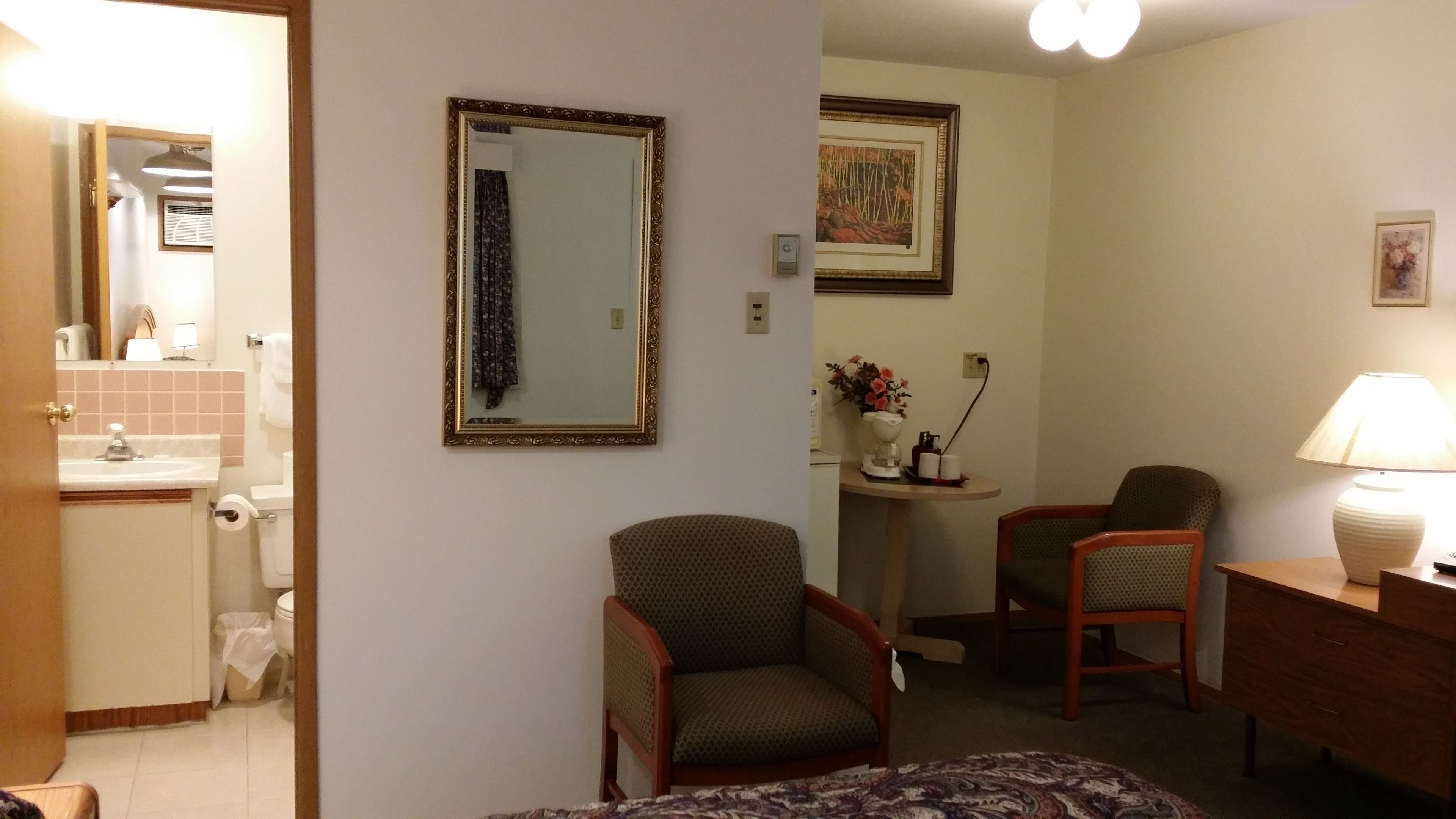 Une chambre d’hôtel comprenant un lit, deux chaises, un bureau, une lampe, un miroir et une salle de bain avec un lavabo et des toilettes.