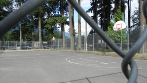 An outdoor basketball court with a chain-link fence, trees, and a clear sky.