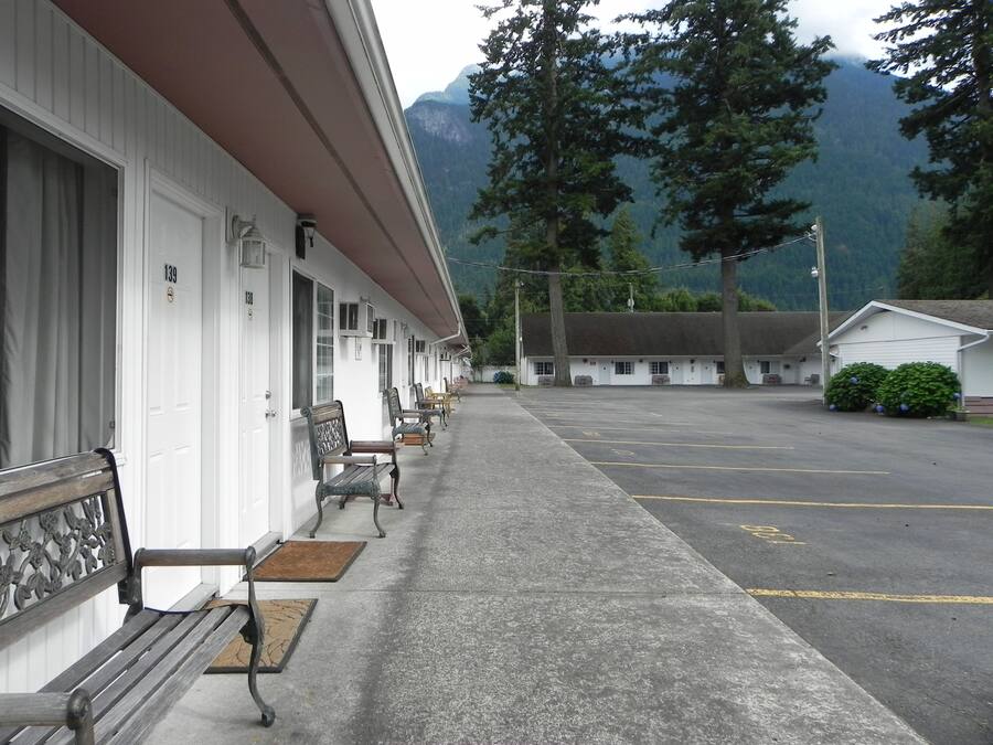 Un motel con una fila de habitaciones individuales, zona de estacionamiento techada y montañas al fondo.