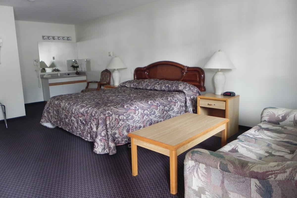 A hotel room with a bed, a desk with a chair, a nightstand, a lamp, and a sofa.
