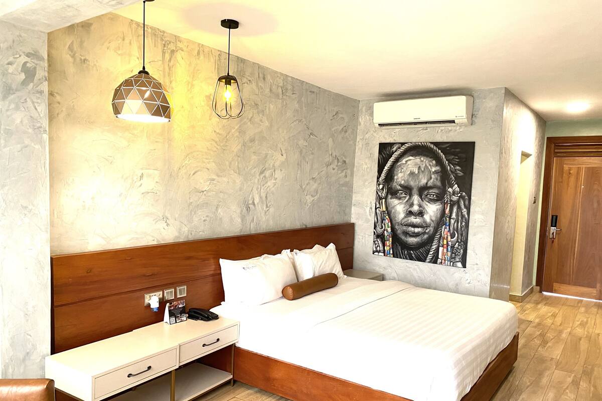 A bedroom with a wooden bed, bedside table, and a desk with a chair. There is a wall-mounted air conditioner and a painting on the wall.