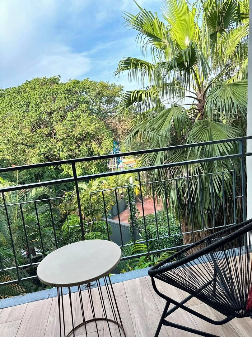 Ein Balkon mit Tisch und Stuhl, der einen üppigen Garten mit verschiedenen Bäumen und einer Palme blickt.