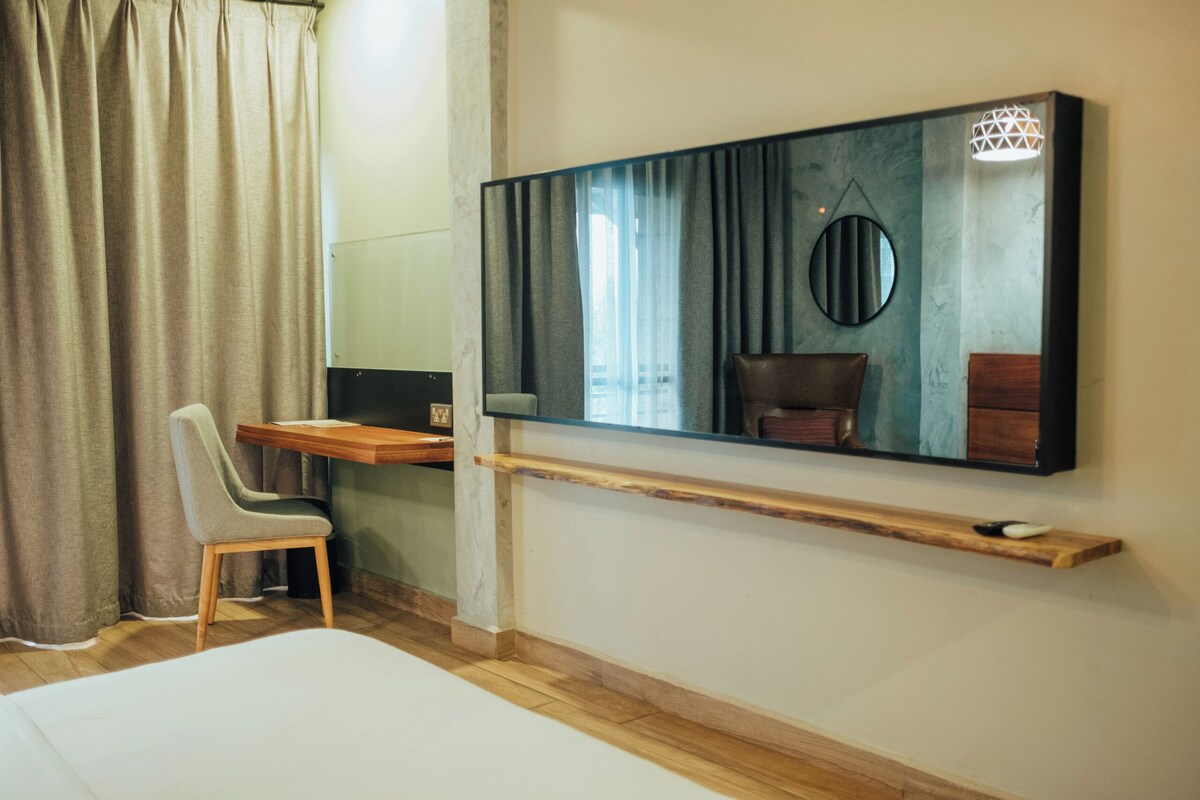 A hotel room with a bed, a desk with a chair, a large mirror, and a shelf with a remote control.