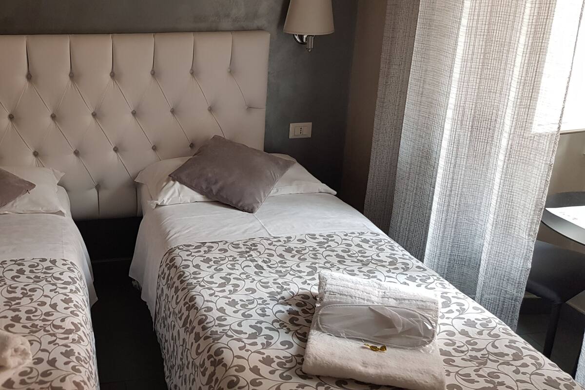 A hotel room with two single beds, each with a patterned bedspread and pillows. There is a large tufted headboard, a wall-mounted lamp, and a window with curtains.
