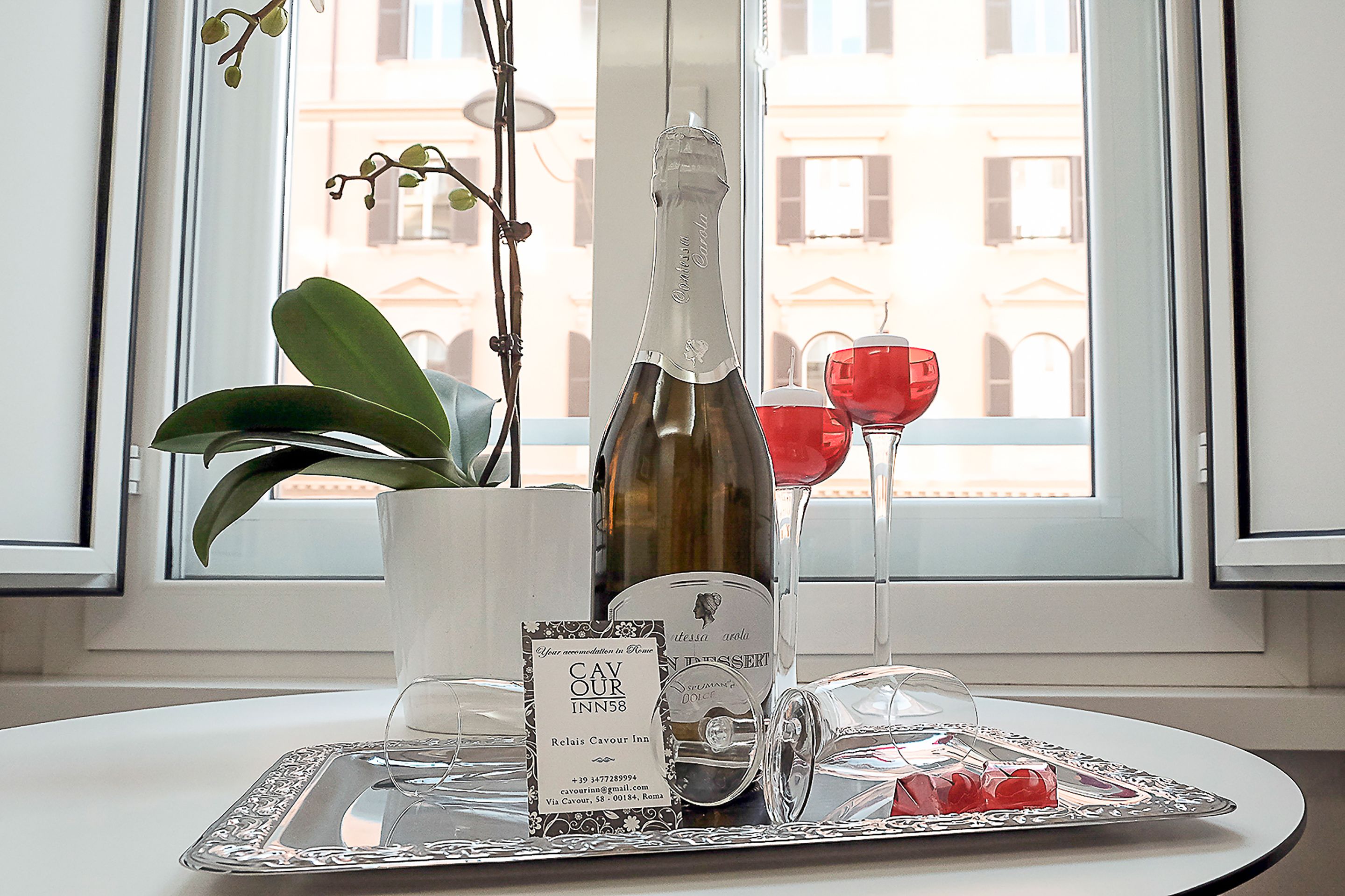 A table with a bottle of wine, a glass, a tray with a dessert, and a card on a white table near a window with a view of buildings.