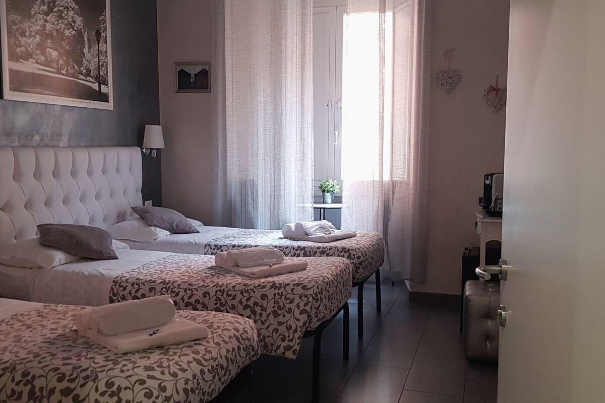 A hotel room with two beds, a large headboard, a bedside table with a lamp, a window with sheer curtains, and a small potted plant.