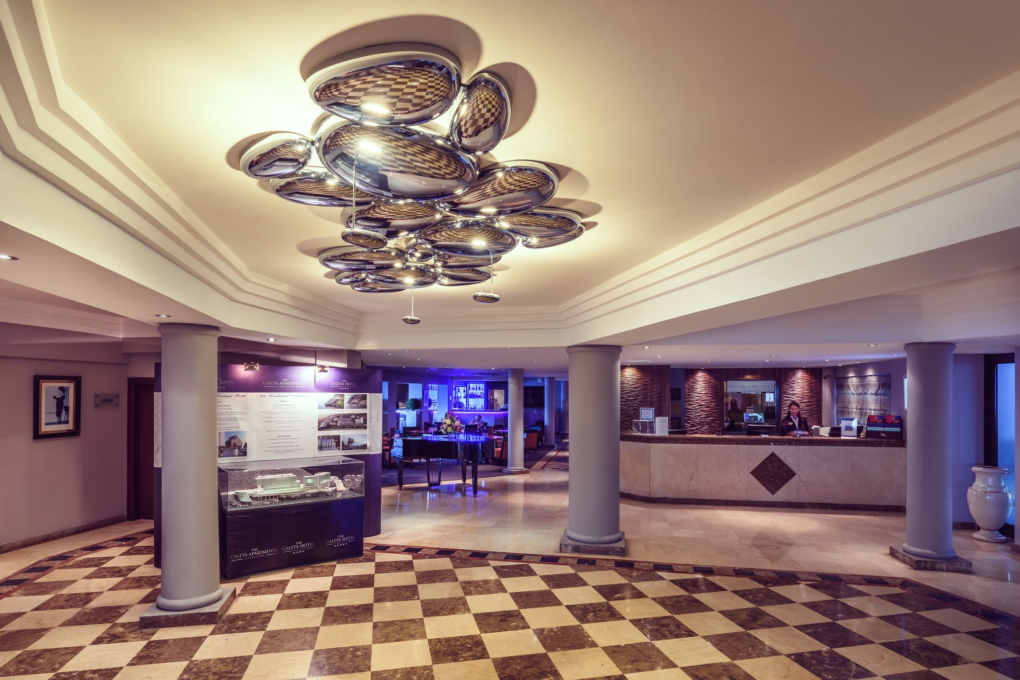 Eine Lobby mit Schachbrettmuster-Boden, einem zentralen Chandelier, einem Empfangsbereich und einer Vitrine mit Informationen.