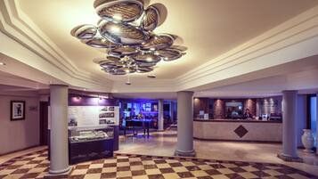 Eine Lobby mit Schachbrettmuster-Boden, einem zentralen Chandelier, einem Empfangsbereich und einer Vitrine mit Informationen.