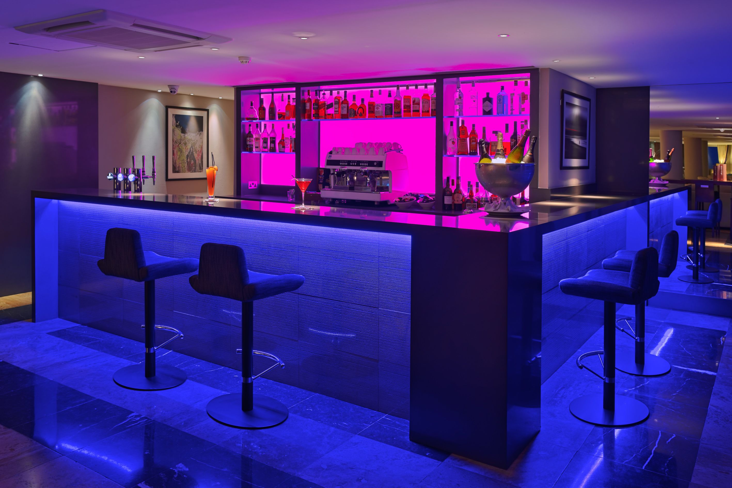 Zona de bar con mostrador iluminado con neón, varias opciones de asientos y una variedad de botellas y vasos en estantes.
