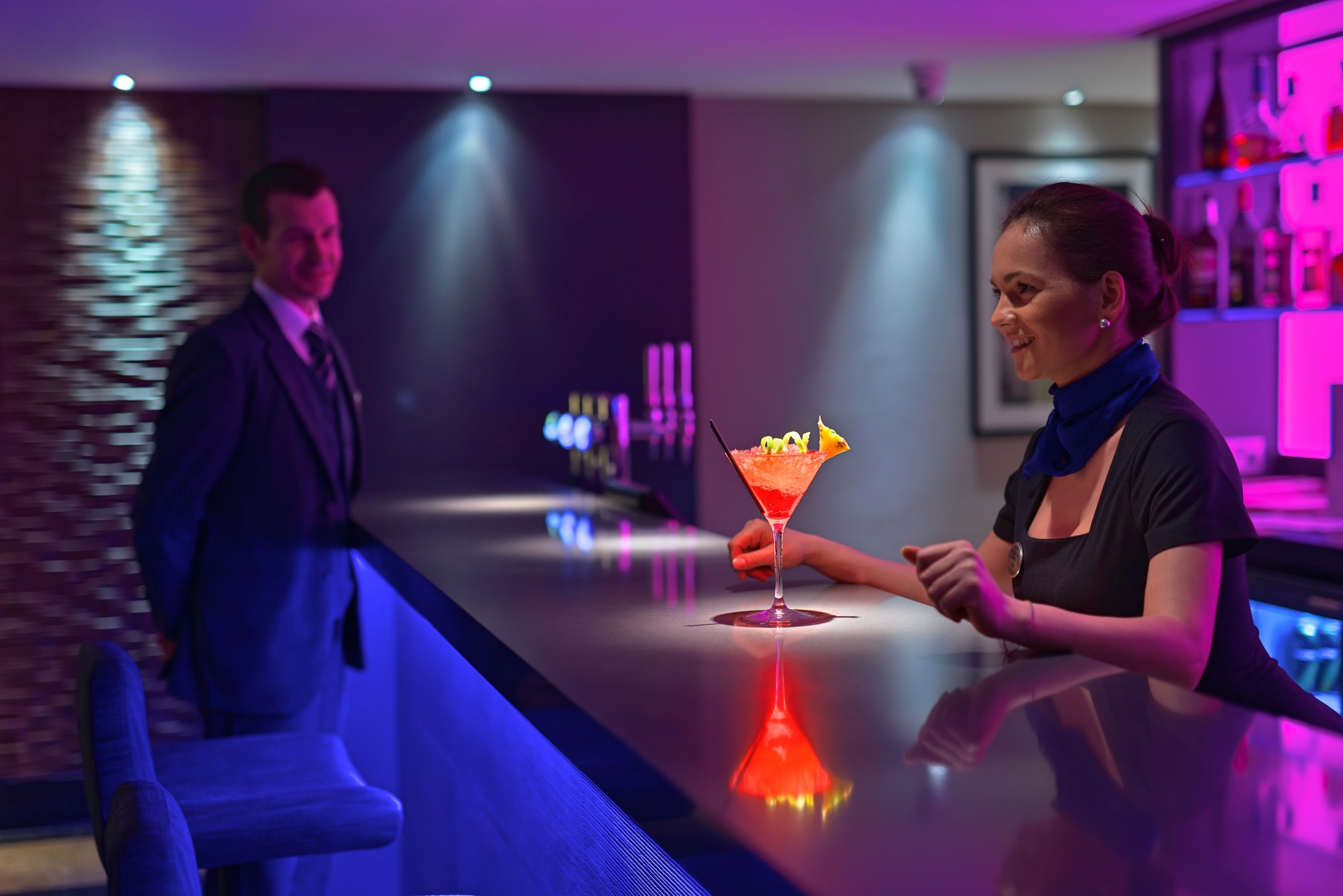 Una zona de bar con mostrador reflectante, una copa de cóctel y una persona sentada en la barra.