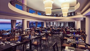 A spacious hotel dining area with multiple tables and chairs, a curved balcony with a railing, and large hanging light fixtures. - The Caleta Hotel Health, Beauty & Conference Centre (Gibraltar)