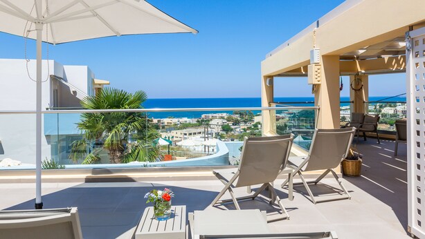 A balcony with two lounge chairs, a table with a vase of flowers, and a view of the sea and cityscape. - Radamanthy's Apartments (Rethymno)