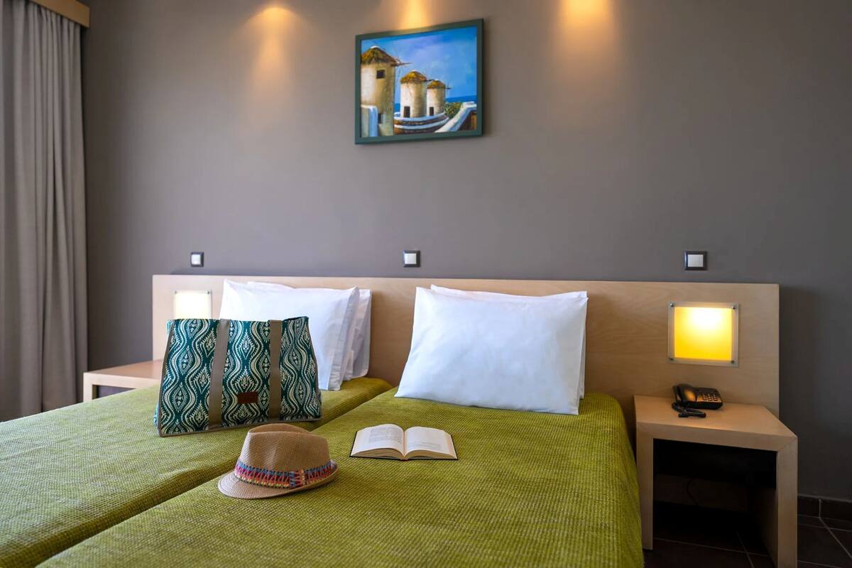 A hotel room with a bed, pillows, a hat, an open book, a painting on the wall, and a bedside lamp.