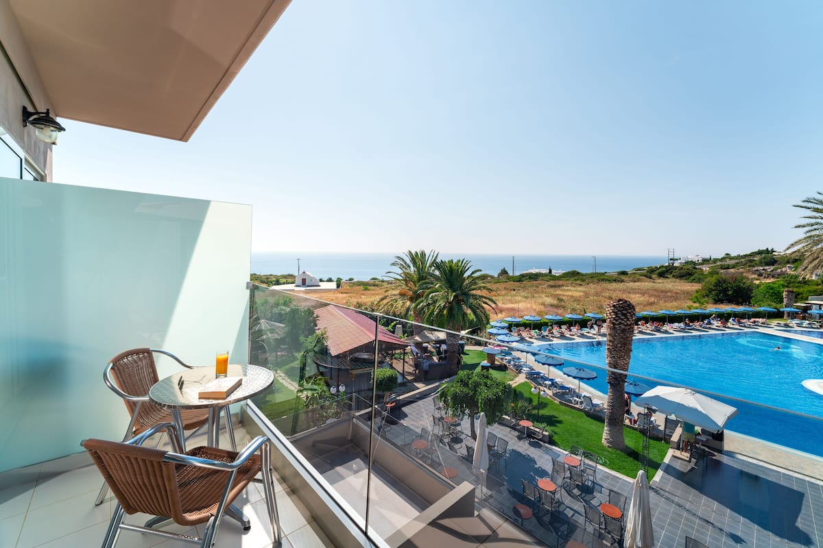 A balcony with a glass table and chairs, overlooking a swimming pool and beach.