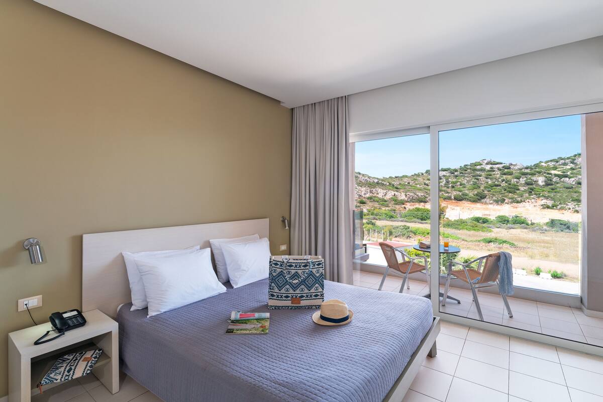 A hotel room with a bed, a bedside table with a phone, a window with a view of the landscape, and a small balcony with a table and chairs.