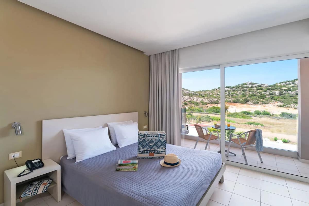 A hotel room with a bed, a bedside table with a phone, a window with a view of the landscape, and a balcony with chairs and a table.