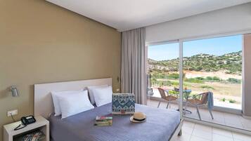 Una habitación de hotel con una cama, una mesita de noche con teléfono, un ventanal con vistas al paisaje y un balcón con sillas y una mesa.