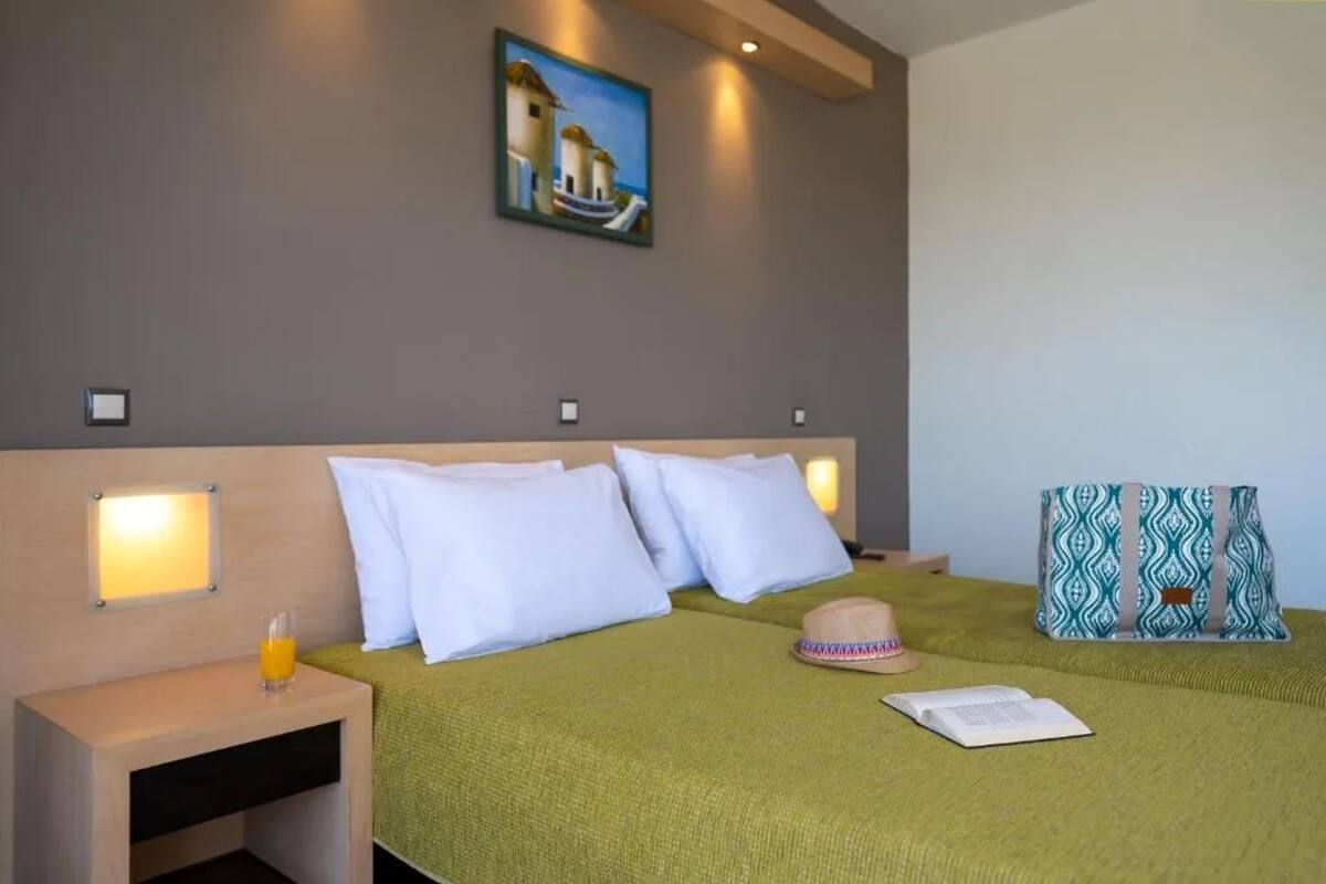 A hotel room with a bed, pillows, a nightstand with a glass of orange juice, a hat, and a patterned bag.
