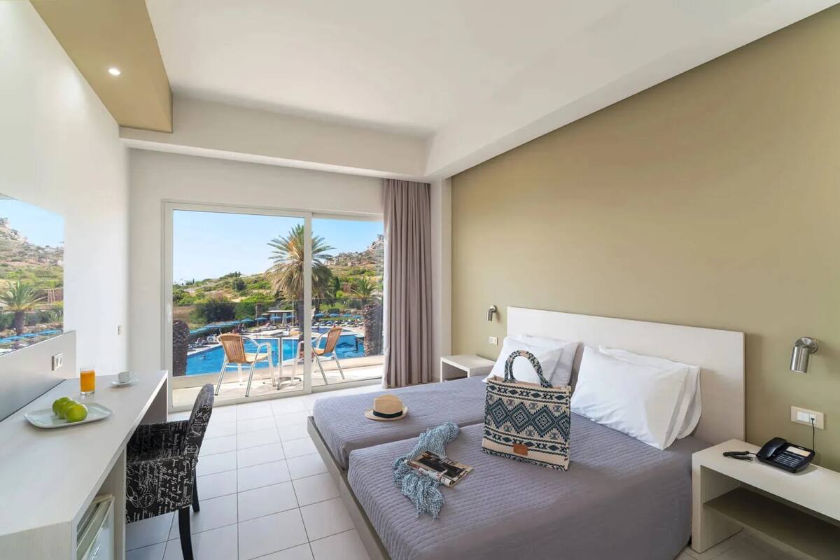 A modern hotel room with a large bed, a desk with a chair, a phone, and a view of a swimming pool and greenery outside.