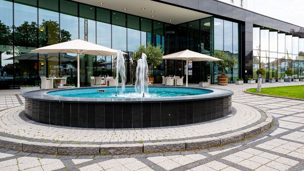 Une fontaine circulaire avec des jets d’eau au centre, entourée d’espaces de détente en plein air et de parasols.