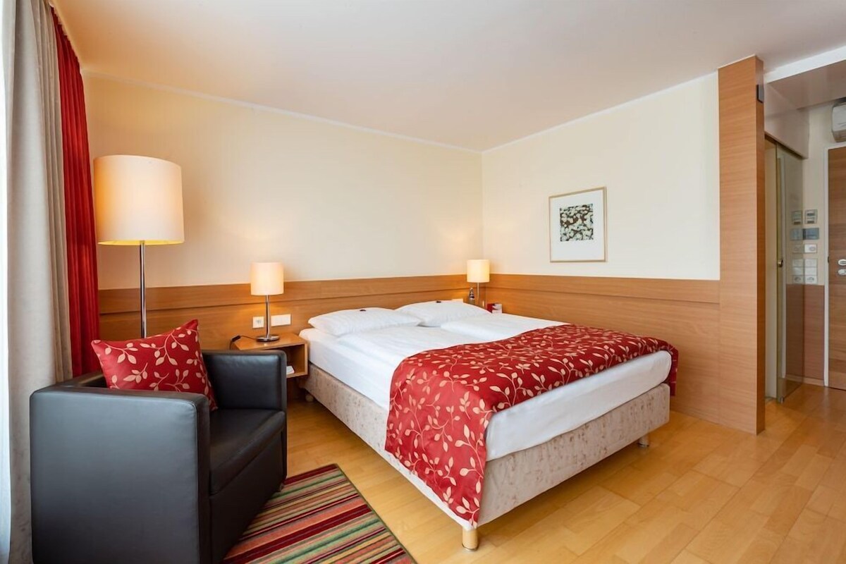 Hotelkamer met een bed, een bank, twee bedlampen, een rode deken met een patroon, een lijst met een kunstwerk aan de muur, en een deur aan de rechterkant.