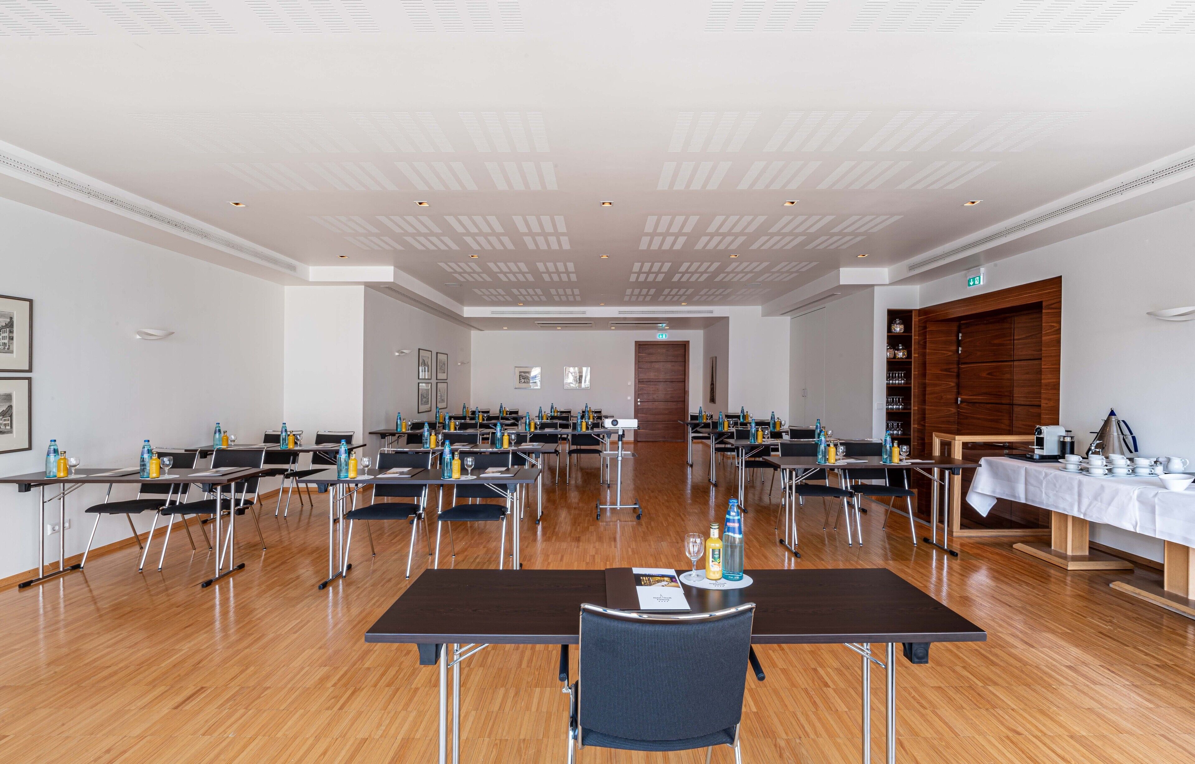 Un salón de conferencias con varias mesas y sillas, piso de madera y techo blanco con iluminación empotrada.