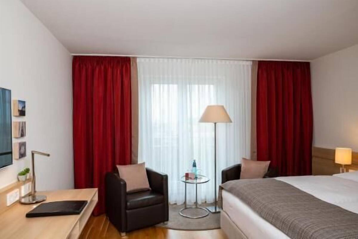 Ein Hotelzimmer mit einem Bett, zwei Stühlen, einem kleinen Tisch, einer Lampe und einem Fenster mit Vorhängen.