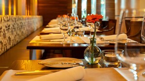 Une salle de restaurant avec une longue table dressée pour le dîner, où les assiettes, les verres et un vase avec une fleur sont disposés avec soin.